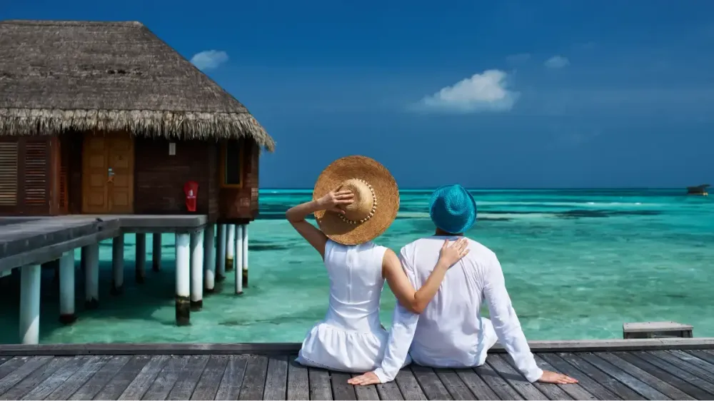 An affluent couple sitting on a wooden pier next to a secluded overwater luxury resort villa, looking out over a turquoise lagoon while enjoying a romantic luxury honeymoon holiday in the Maldives.